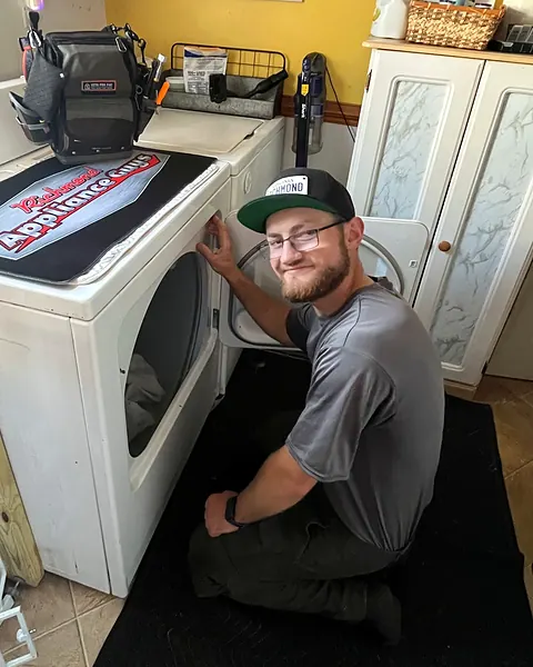 Viktor kneeling at a dryer and smiling at the camera, tool bag on the machine beside him